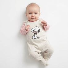 Load image into Gallery viewer, A baby lying on their back wearing a cream-colored footed jumpsuit with a Snoopy graphic holding flowers, layered over a light pink long-sleeve shirt, against a plain white background.
